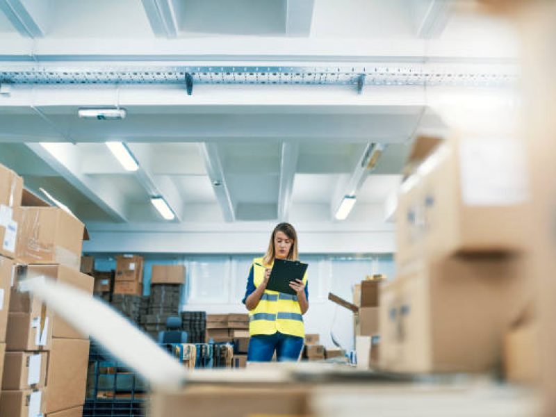 Photo of Young woman with clipboard at logistics company warehouse. Worker In Warehouse Preparing Goods For Dispatch. Business woman is showing a clipboard in hand and checking boxes